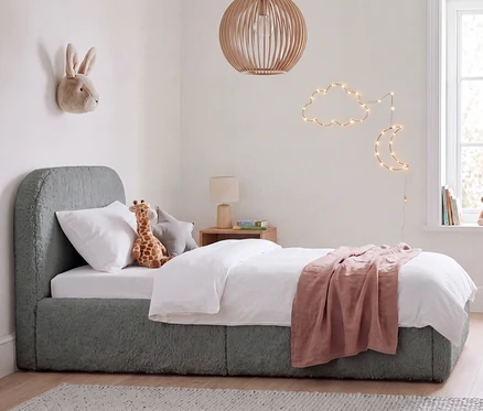 Kinderzimmer mit einem grauen Bett mit Teddy-Stoff, weißer Bettwäsche und einer rosa Decke. Ein Stofftier-Hase hängt an der Wand, eine Giraffe liegt auf dem Bett. Eine Wolken- und Mond-Lichterkette schmückt die Wand, und eine Holzlampe hängt von der Decke.