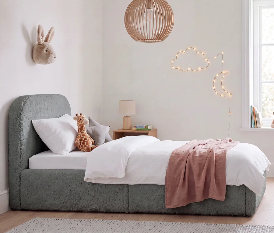 Kinderzimmer mit einem grauen Bett mit Teddy-Stoff, weißer Bettwäsche und einer rosa Decke. Ein Stofftier-Hase hängt an der Wand, eine Giraffe liegt auf dem Bett. Eine Wolken- und Mond-Lichterkette schmückt die Wand, und eine Holzlampe hängt von der Decke.