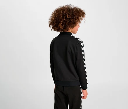 Person von hinten, mit lockigem Haar. Trägt eine schwarze hummel Archive Reg Poly Zip Jacket mit weißen Chevron-Streifen an den Ärmeln und schwarze Hose mit weißen Streifen.