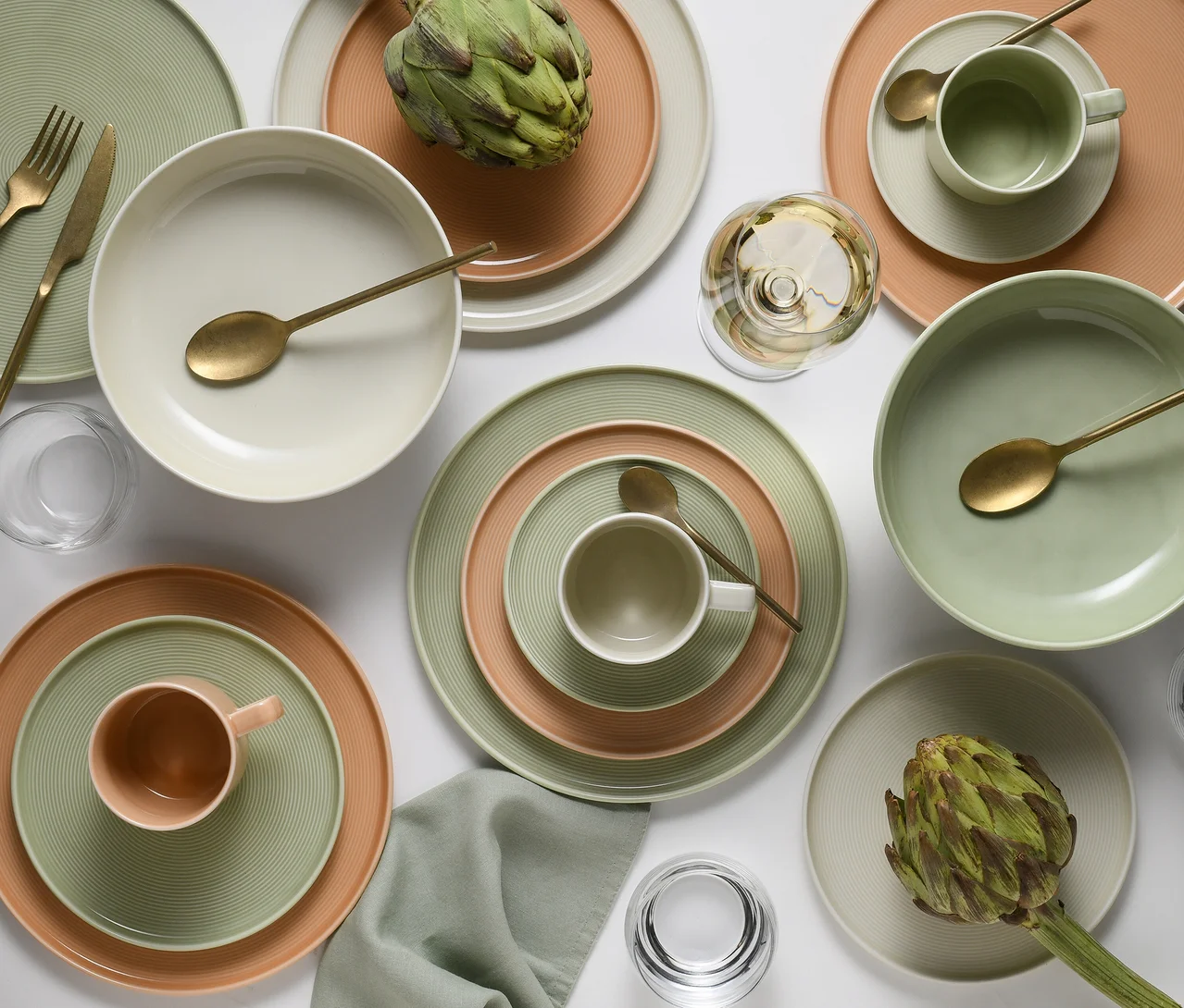 Das creaTable Service »Session Romance« mit Tellern, Schalen und Tassen in Grün, Pfirsich und Cremeweiß, dazu goldfarbenes Besteck, Artischocken und Gläser auf weißem Untergrund.