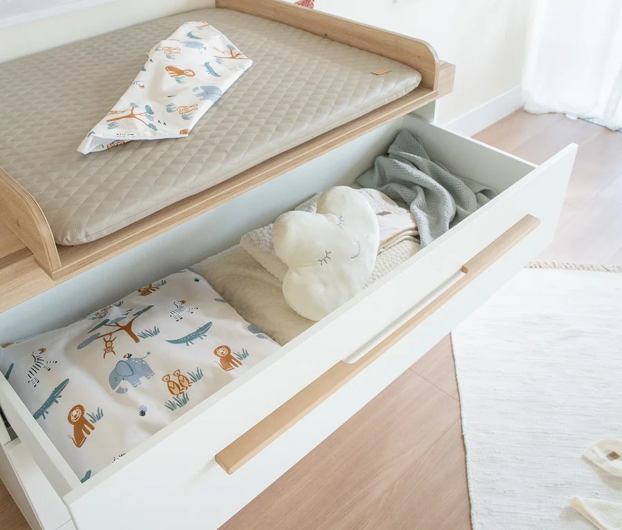 Weißer Wickeltisch des Roba 3-teiligen Kinderzimmer-Sets »Lumo« mit hellbrauner Wickelauflage und einem Tuch mit Tiermotiven. Eine offene Schublade zeigt ein Herz-Kissen, eine Decke und Bettwäsche mit Tiermustern.