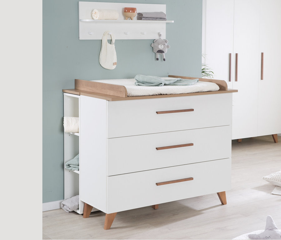 Ein Kinderzimmer mit einem weißen Wickeltisch mit drei Schubladen und Holzdetails, einem weißen Regal und einem weißen Schrank.
