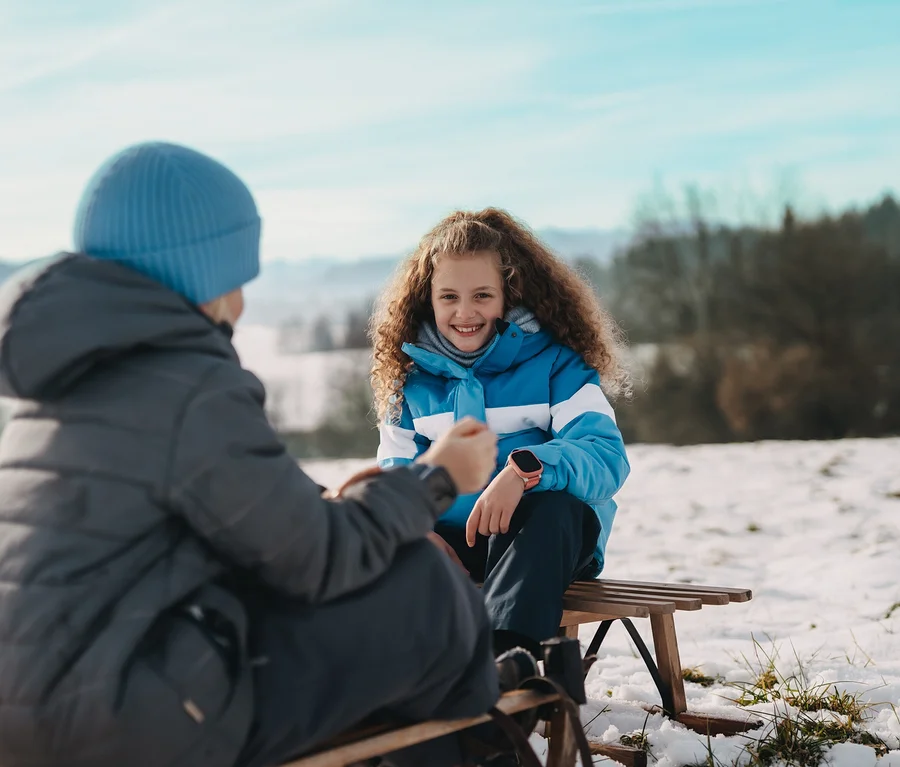 Ein lächelndes Mädchen mit lockigem Haar sitzt auf einem Holzschlitten im Schnee und trägt eine pinke Xplora X6 Play Kids Watch am Handgelenk, während ein anderes Kind mit blauem Beanie ihr gegenüber sitzt.