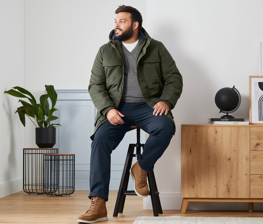 Ein Mann sitzt auf einem Hocker und trägt eine olivgrüne Parka, einen grauen Merino-Pullover mit V-Ausschnitt, ein weißes T-Shirt und Veloursleder-Boots.