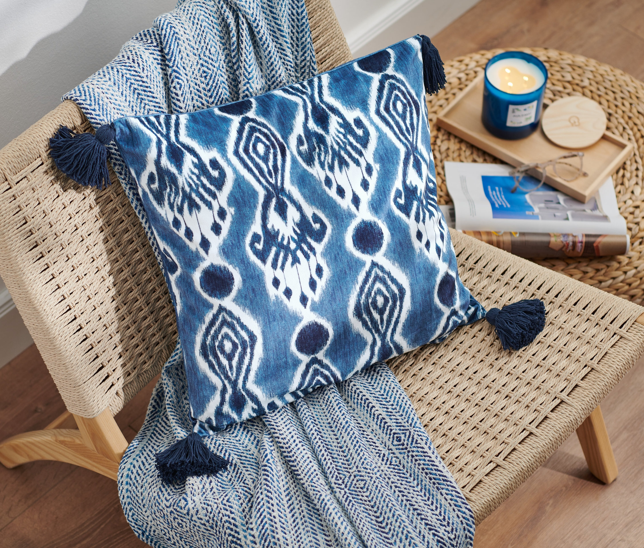 Auf einem Korbsessel liegen ein blaues Samt-Dekokissen-Bezug mit Quasten und eine blaue Decke. Neben dem Sessel steht ein Korbtisch mit einer blauen Kerze und einer Zeitschrift.