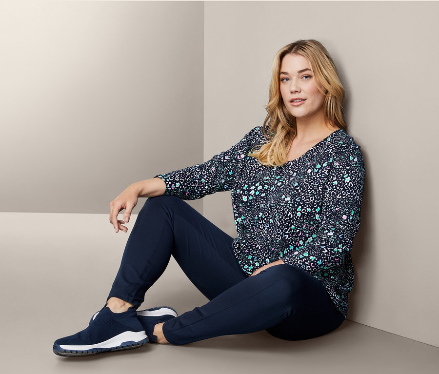 Frau sitzt auf dem Boden und trägt Slip-on-Sneaker, Langarmshirt und Bodyforming-Jerseyhose.