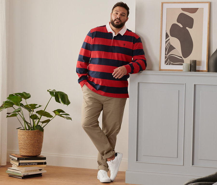 Mann lehnt an Wand, trägt Sneaker, camelfarbene Chino und navyfarben-rotes Langarm-Poloshirt.