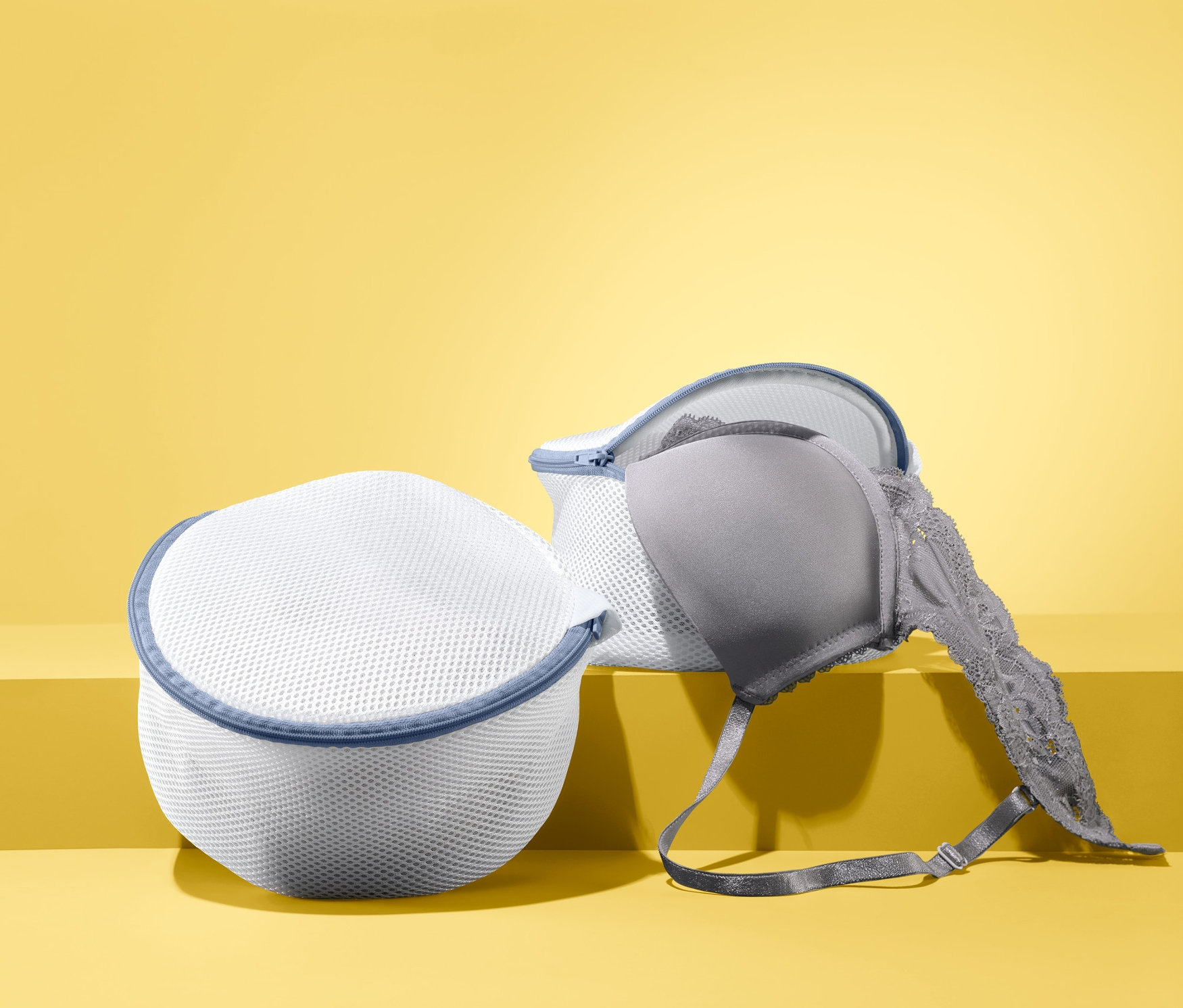 Zwei BH-Wäschenetze liegen vor gelbem Hintergrund, in einem davon befindet sich ein grauer BH.