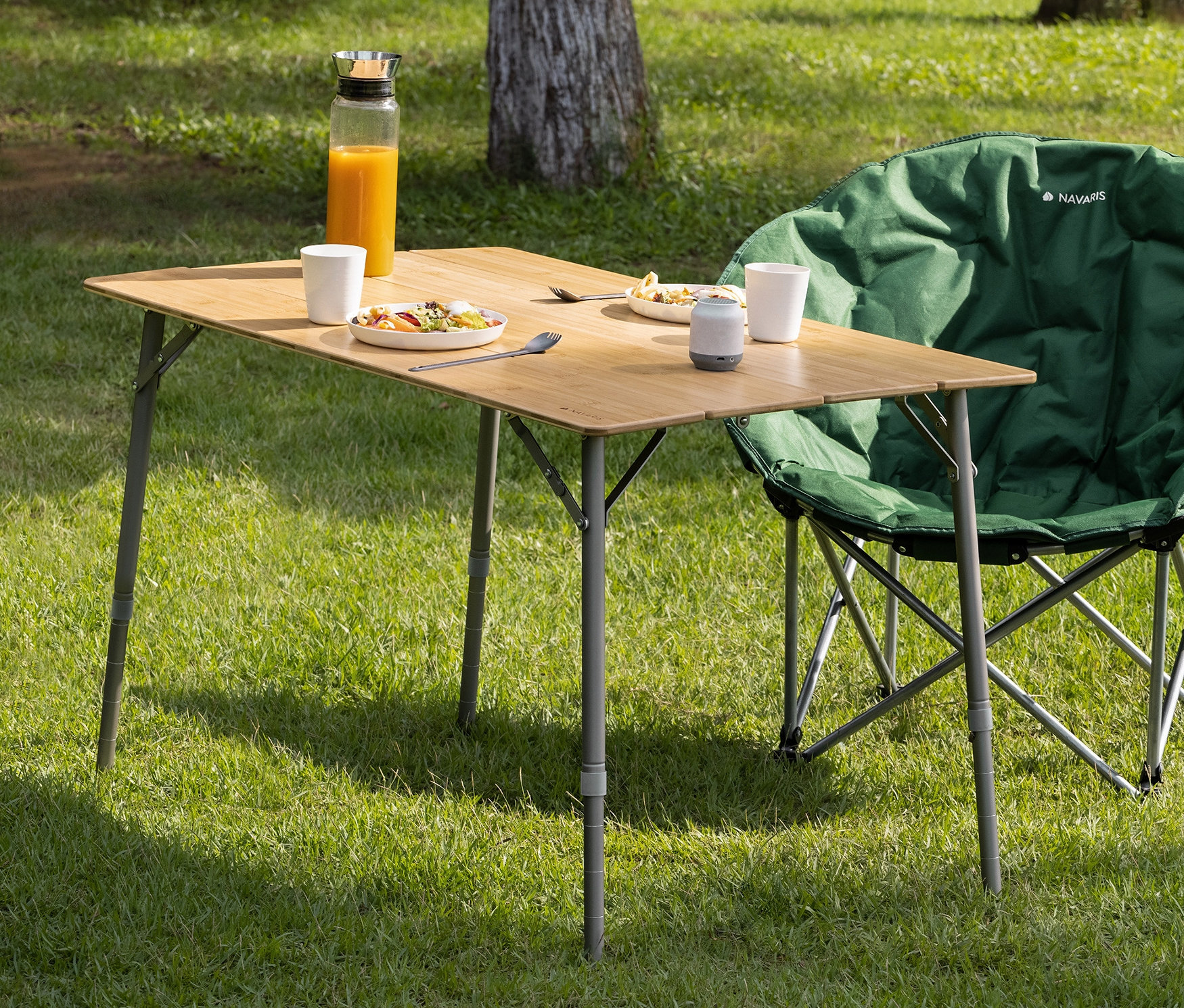 Navaris Bambus-Campingtisch mit Speisen, Getränken und einem grünen Campingstuhl auf einer Wiese.