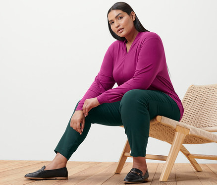 Frau sitzt auf einem Stuhl, trägt ein berryfarbenes Langarmshirt mit V-Ausschnitt und eine Stretchhose.