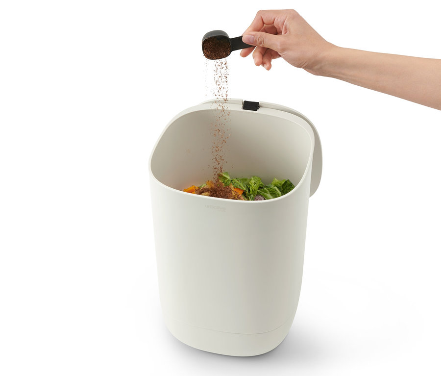 Eine Hand schüttet mit einem Löffel Pulver in einen Bokashi-Kompostier-Mülleimer, der mit Essensresten gefüllt ist.