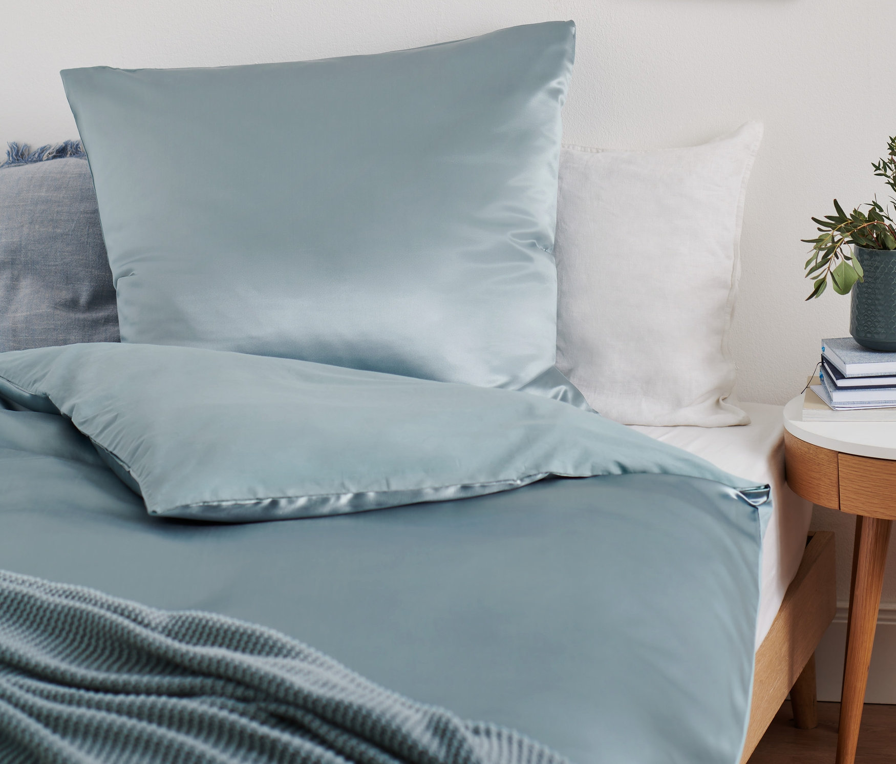 Hellblaue Premium-Seiden-Baumwoll-Bettwäsche in Normalgröße auf einem Bett. Neben dem Bett steht ein Tisch mit einer Topfpflanze.