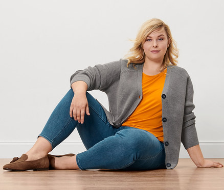 Blonde Frau sitzt auf Holzboden, trägt Treggings »Fit Nelly«, orangefarbenes Blusenshirt, Grobstrick-Cardigan und Loafer aus Veloursleder.