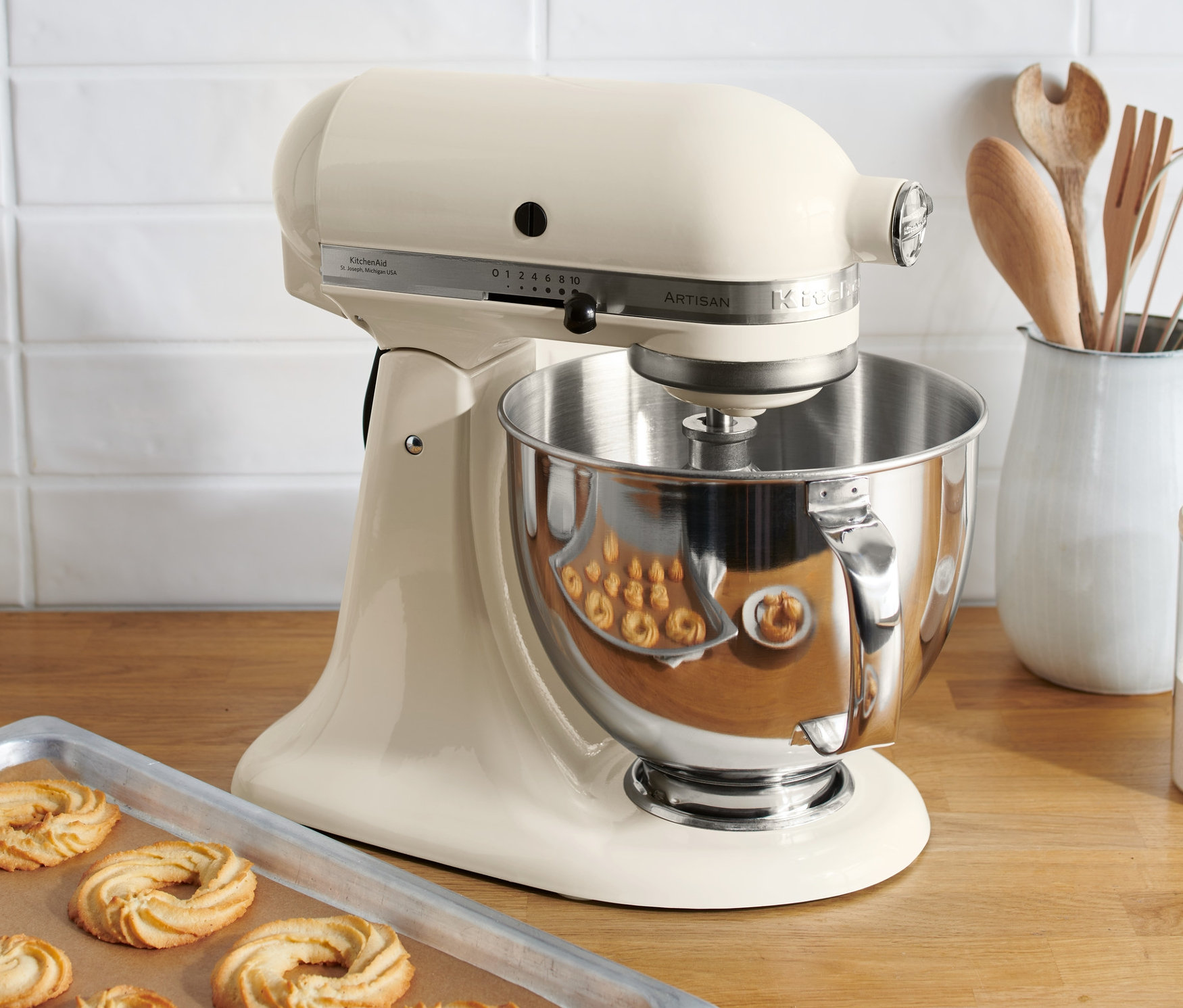 Eine KitchenAid Küchenmaschine »Artisan 5KSM125EAC« in Creme steht auf einer Holzfläche neben einem Backblech mit Keksen.