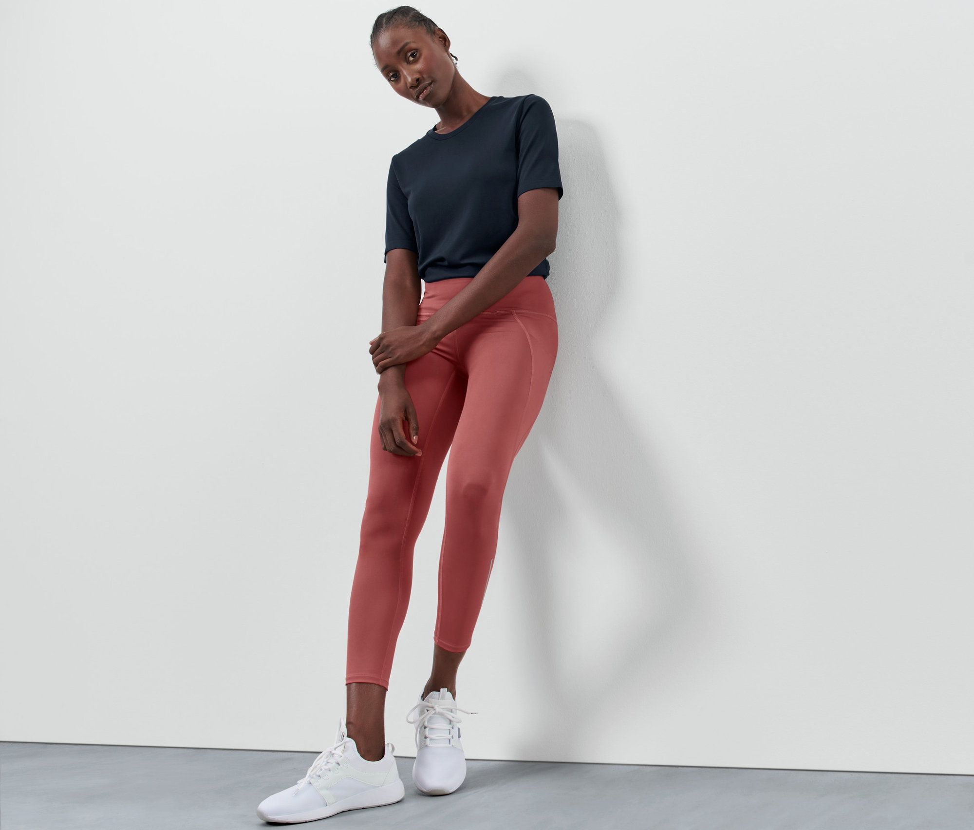 Eine Frau lehnt an einer Wand und trägt eine Sporttight in Terrakotta, ein marineblaues T-Shirt und weiße Turnschuhe.