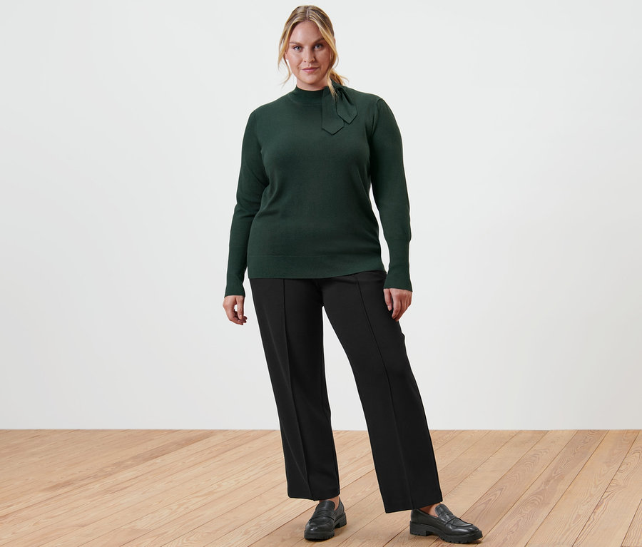 Frau posiert in Feinstrickpullover mit Schleifendetail, schwarzer Jerseyhose mit Biese und Leder-Loafern.