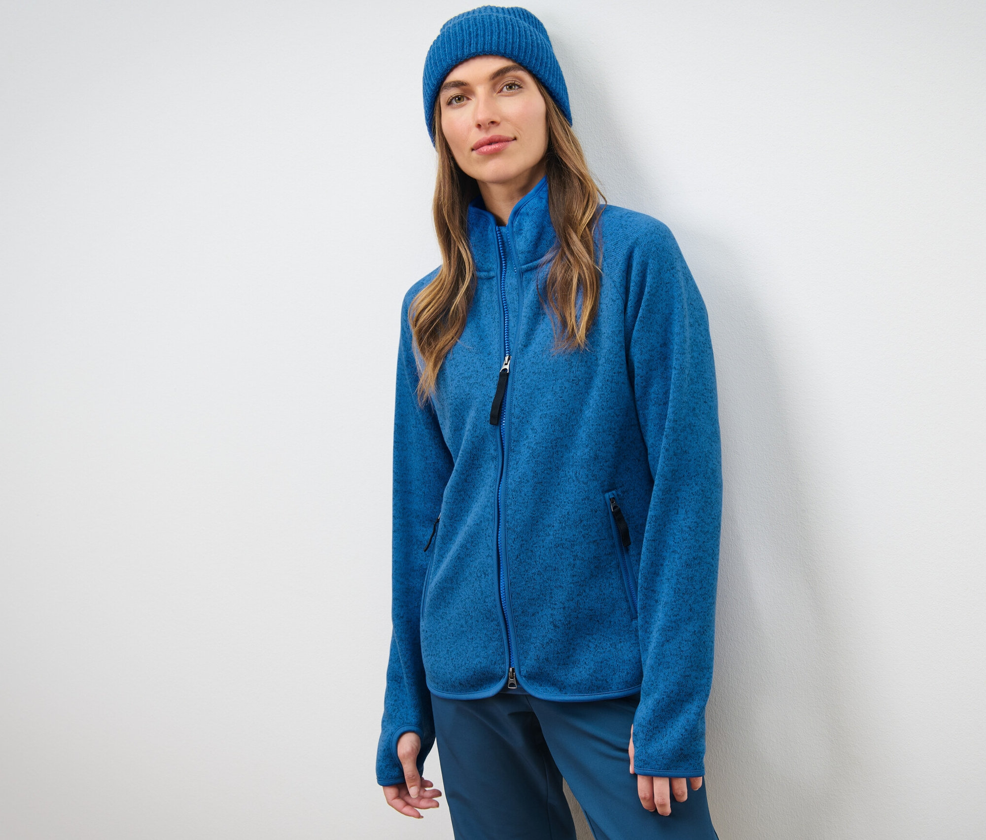Frau vor einer weißen Wand trägt eine blaue Strickfleece-Jacke und eine blaue Mütze.