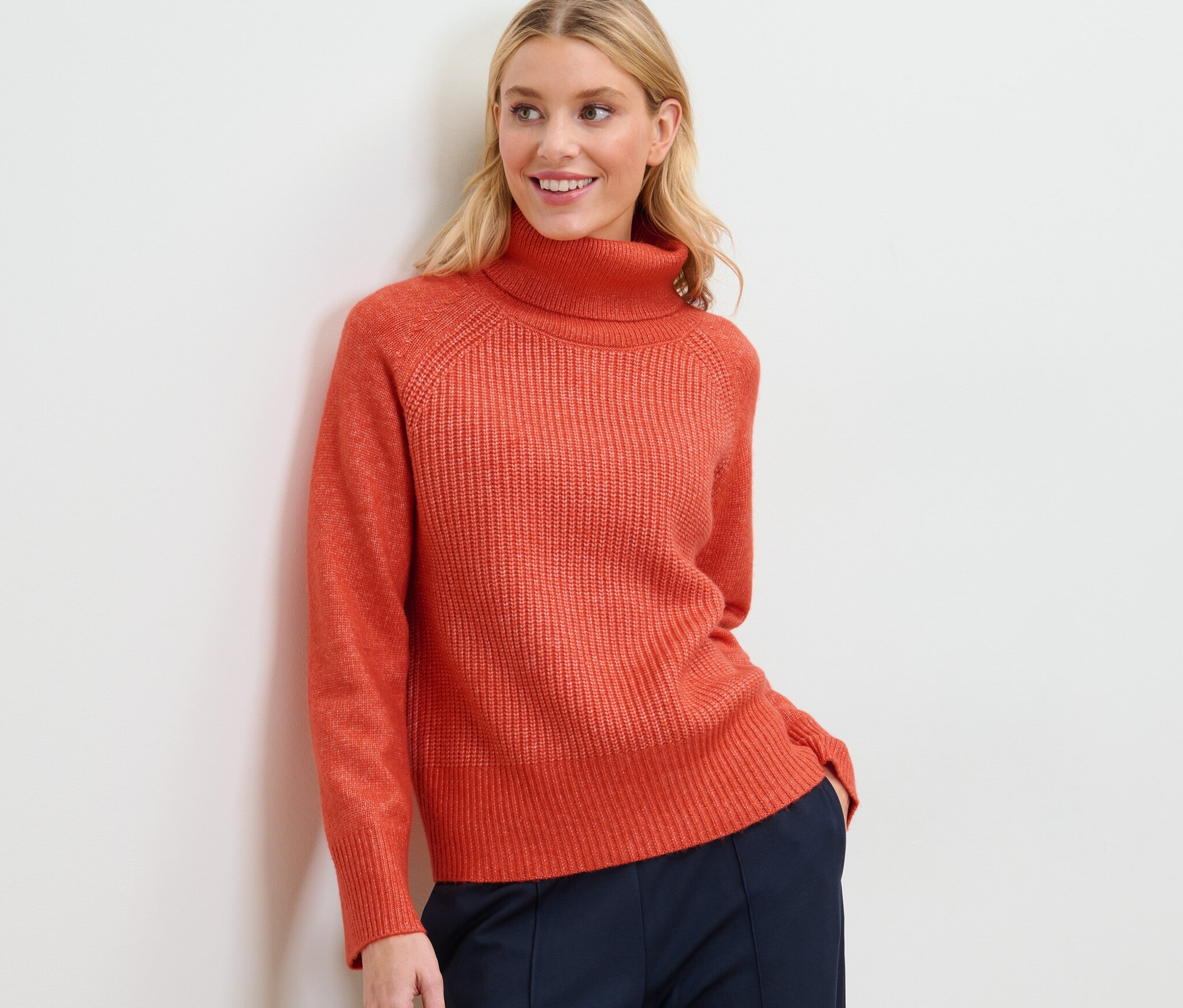 Blonde Frau posiert in orangefarbenem Grobstrickpullover mit Rollkragen und marineblauer Hose.