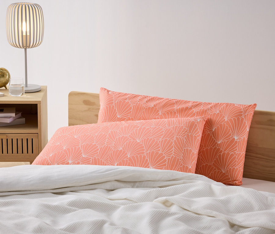 Zwei korallenfarbene Microfaser-Kopfkissenbezüge mit weißem Blumenmuster liegen auf einem Bett. Ein Nachttisch mit Lampe steht neben dem Bett.