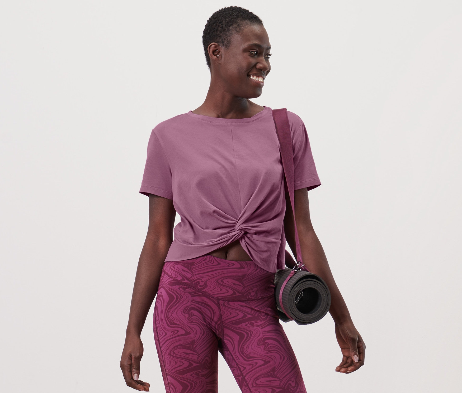 Frau trägt ein Sportshirt und Leggings, mit einer Tasche und einer Yogamatte über der Schulter.