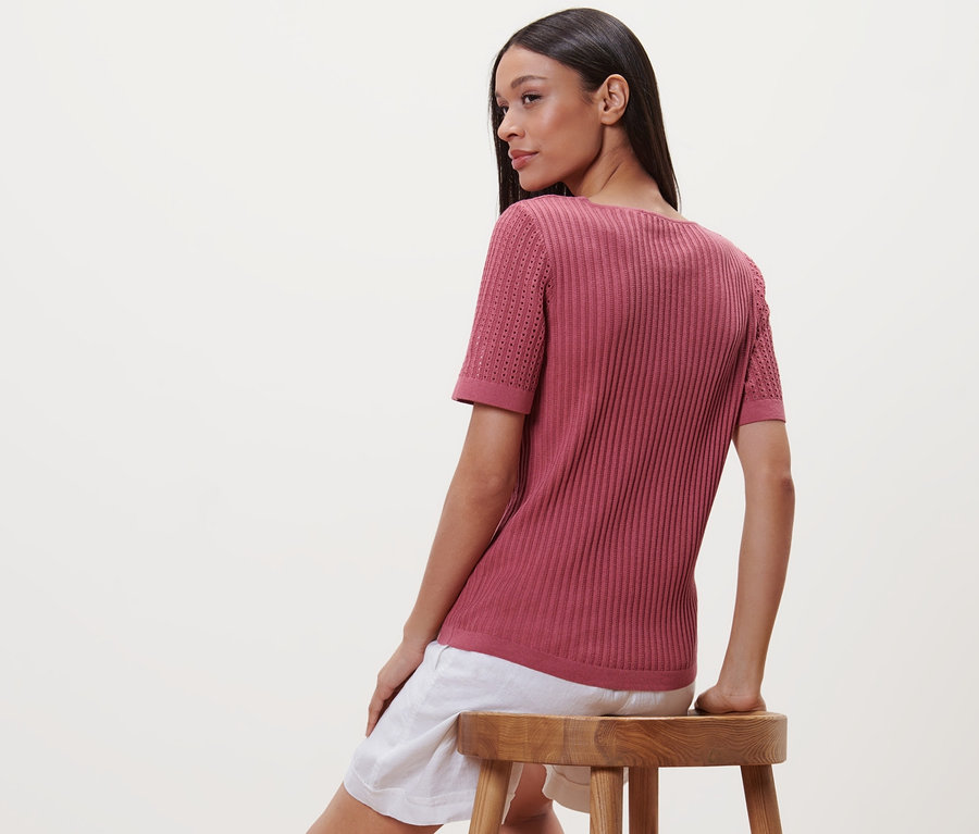 Frau sitzt auf einem Hocker und trägt ein beerefarbenes Strick-Shirt.
