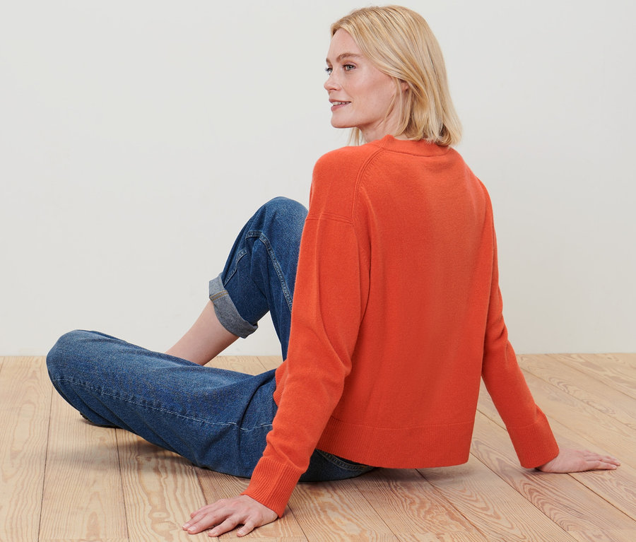 Blonde Frau sitzt auf Holzboden und trägt einen orangefarbenen Feinstrick-Pullover mit Cashmere und Jeans.