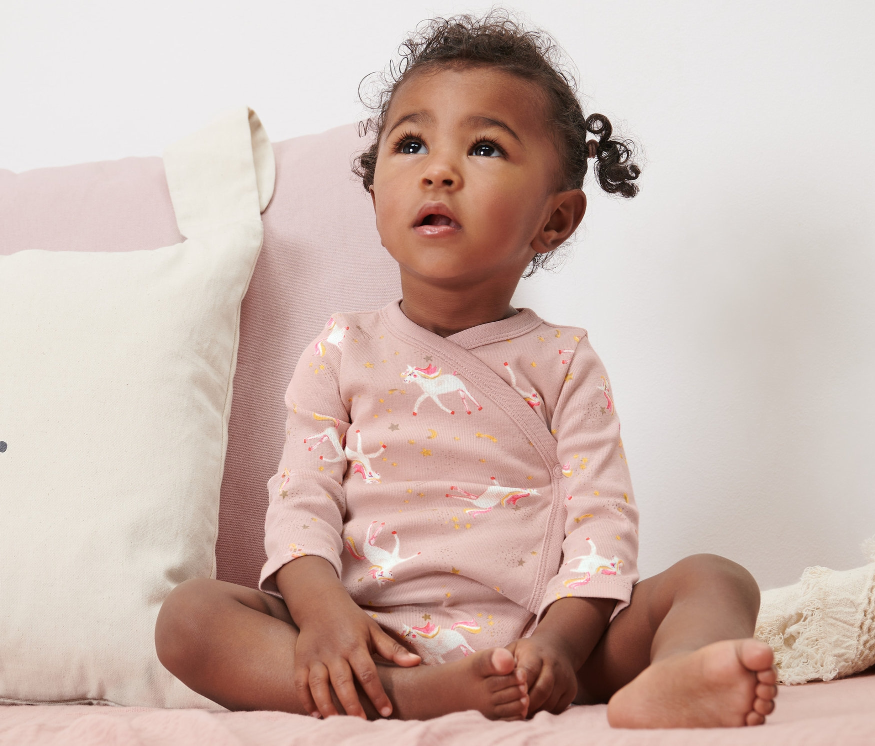 Kleines Kind sitzt auf einem Bett und trägt einen 5er-Pack Baby-Bodys mit Mitwachsfunktion, Einhorn.