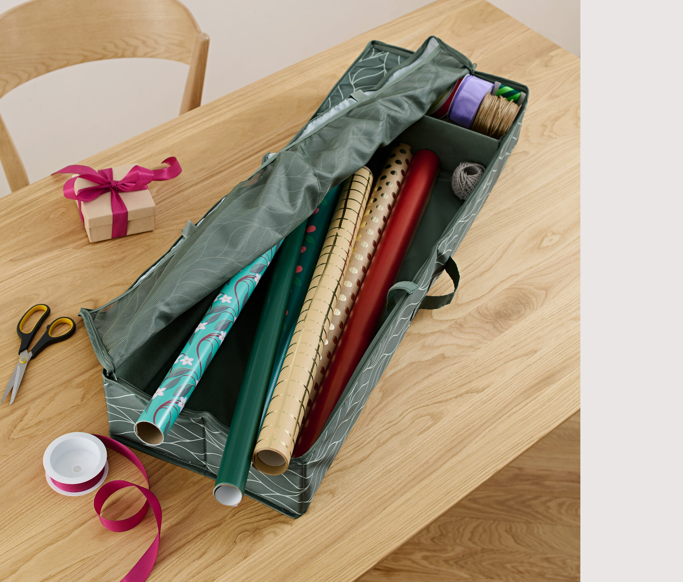 Auf einem Tisch liegen ein Geschenkpapier-Organizer, eine Schere, ein Geschenkband und ein verpacktes Geschenk.
