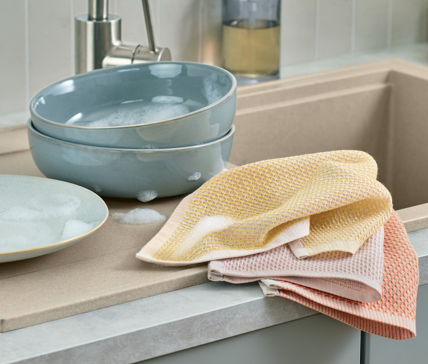 Zwei blaue Schüsseln mit Seifenwasser und ein leerer Teller liegen nebeneinander auf einer Abtropffläche. Daneben liegen 3 Spültücher.