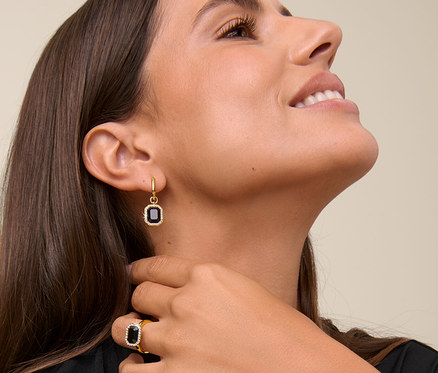 Frau lächelt und berührt ihren Hals, sie trägt eine Kette Royal Black, Creolen Royal Black und einen Ring Royal Black.