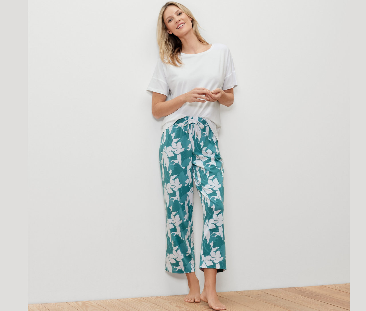 Blonde Frau lehnt an einer weißen Wand und trägt ein weißes T-Shirt und eine grüne Pyjamahose mit Muster.