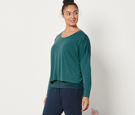 Eine Frau posiert in Yogahose und einem 2-in-1-Sportshirt, dunkelsmaragdgrün. Daneben liegen eine zusammengerollte Yogamatte und eine Wasserflasche.
