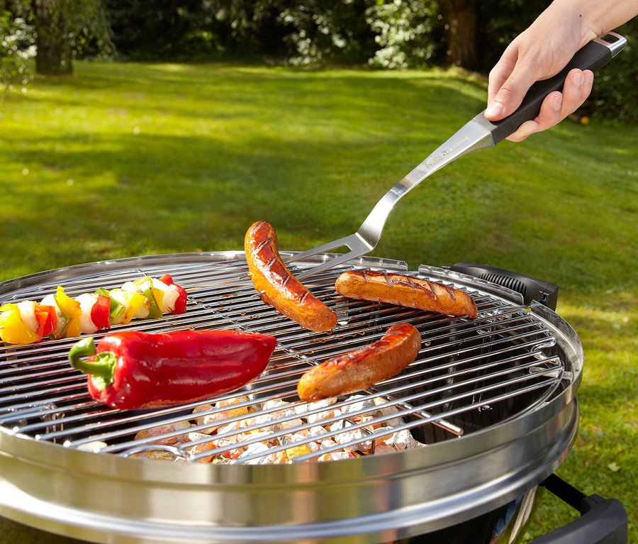 Würste, eine rote Paprika und ein Gemüsespieß liegen auf dem Grill. Eine Hand wendet mit Landmann Grillzubehör eine Wurst.