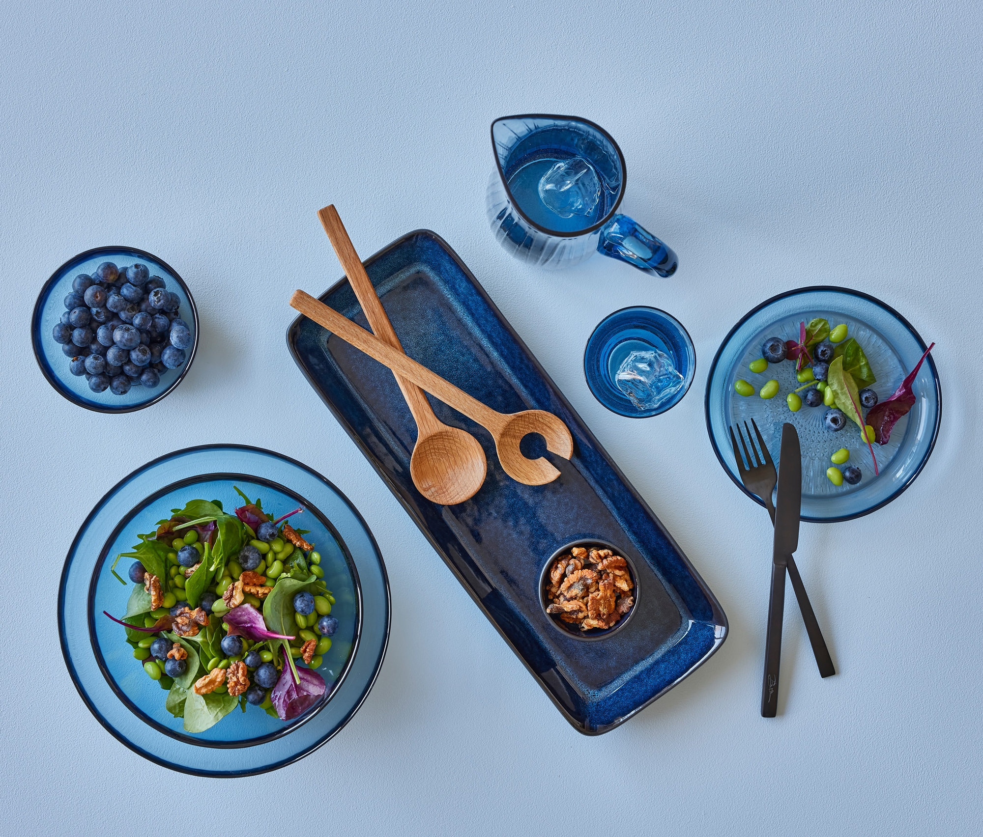 Eine blaue Bitz Salatschüssel »Kusintha« mit Obstsalat und Blaubeeren, zwei Bitz Glasteller »Kusintha« und zwei Bitz Glasschalen »Kusintha« auf hellblauem Grund.