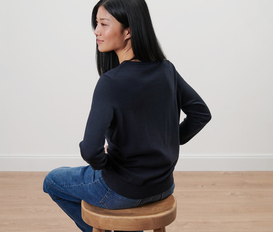 Frau sitzt auf einem Holzhocker und trägt einen dunkelblauen Merino-Feinstrickpullover.