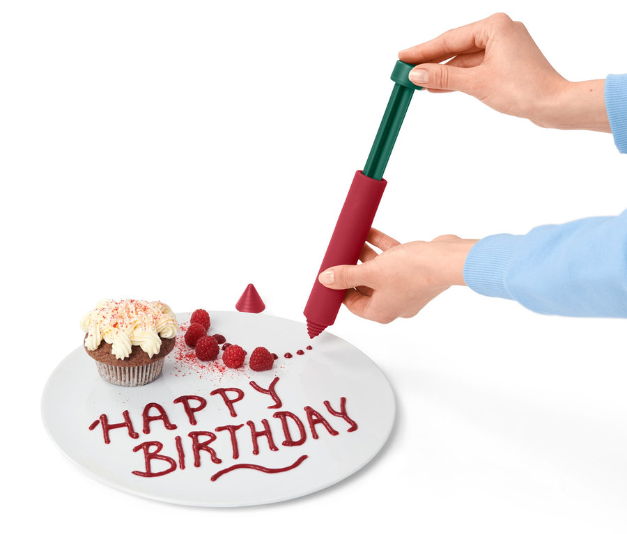 Frau verziert Teller mit "Happy Birthday" mit Dekorierstift, daneben Muffin und Himbeeren.