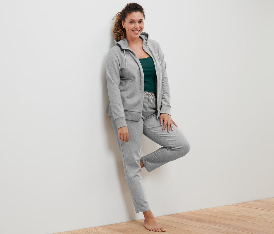 Eine Frau lehnt an einer Wand und trägt eine graue Sweatjacke, zwei Tops in Grün und Offwhite und eine graue Sweathose.