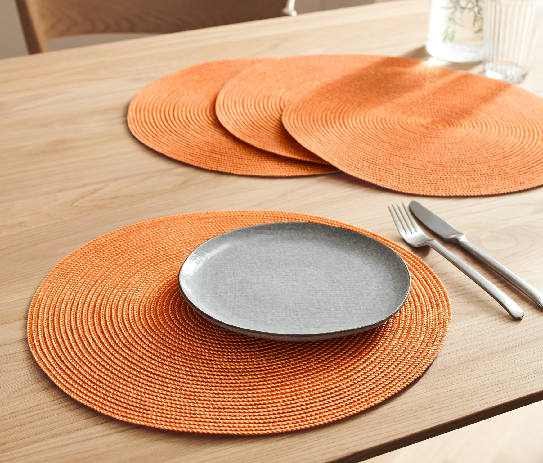 Auf einem Tisch liegen 4 orangefarbene Platzsets. Auf einem davon liegen ein grauer Teller, ein Messer und eine Gabel.