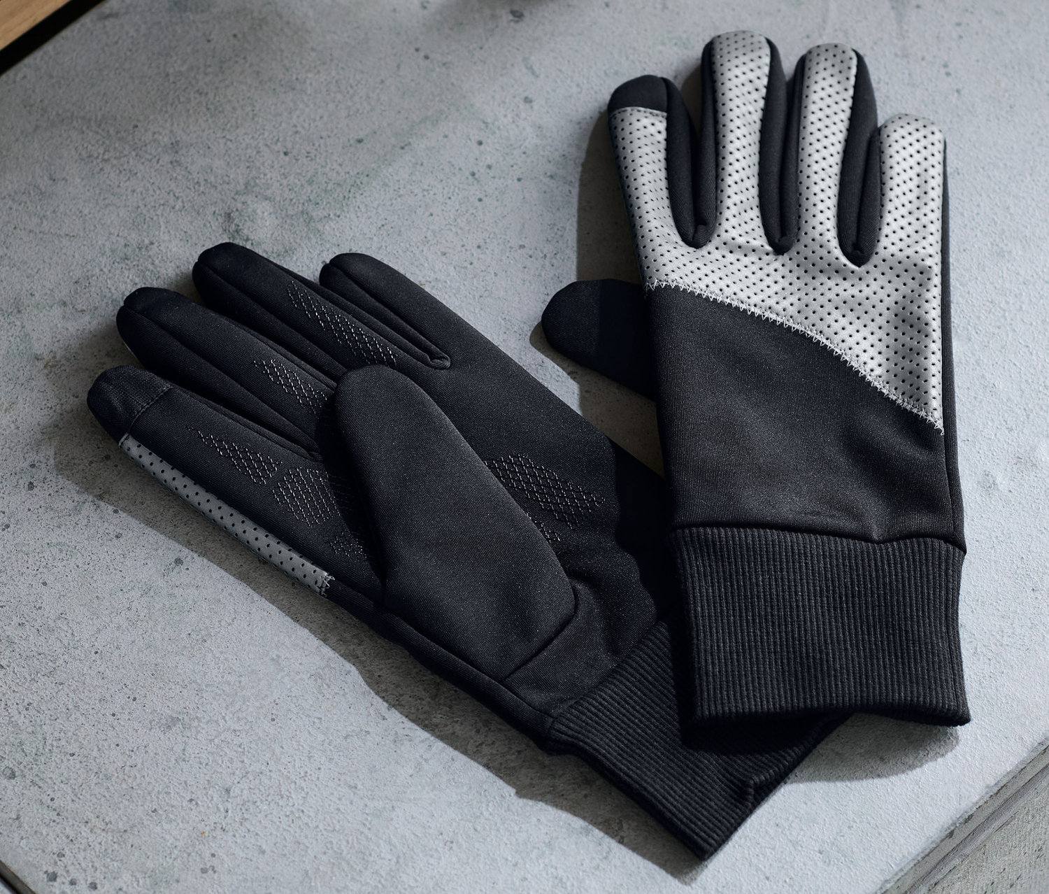 Zwei Paar Windprotection-Handschuhe liegen auf einer grauen Oberfläche neben Ohrhörern und einer Wasserflasche.