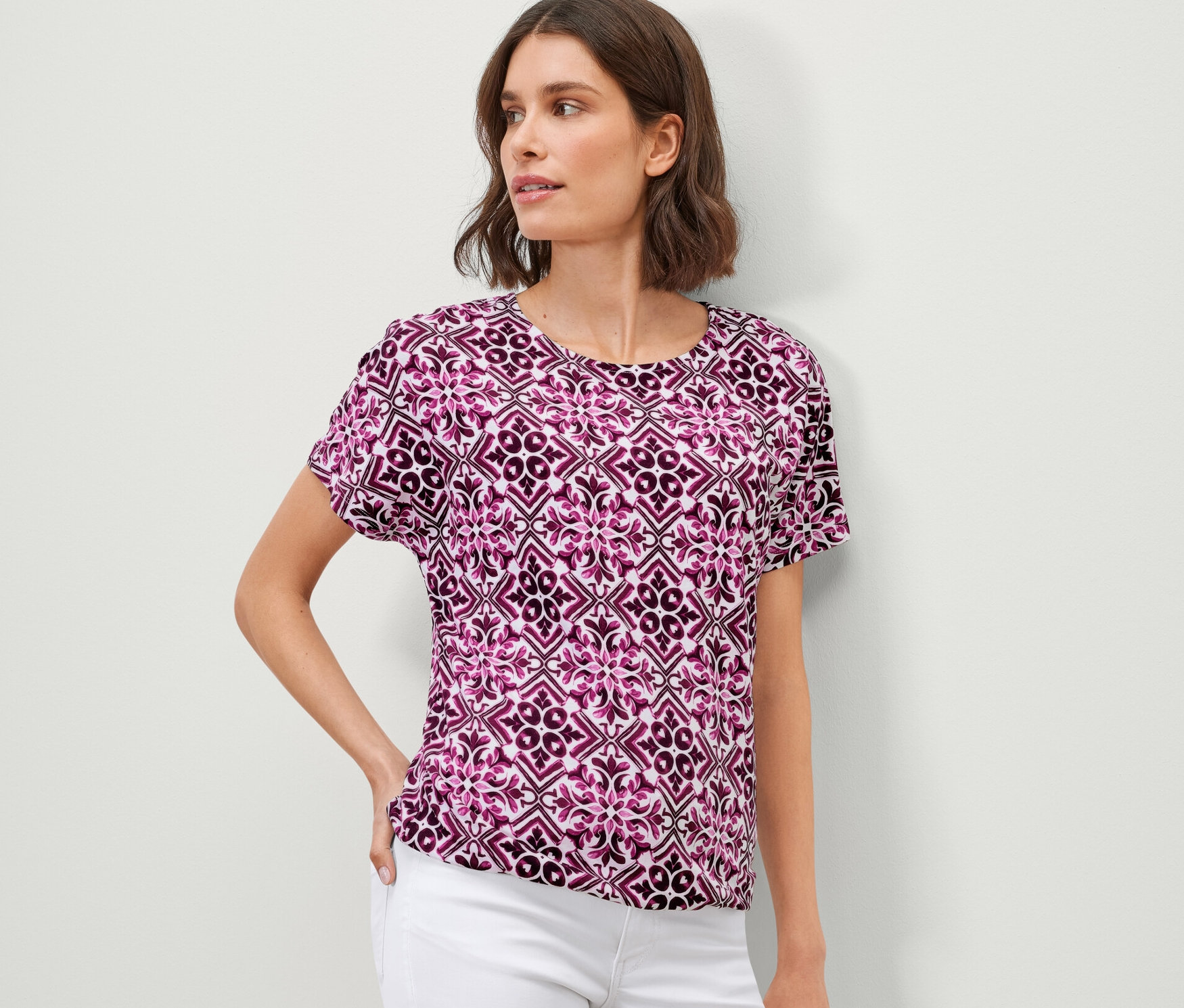 Frau posiert in einem bedruckten Blusenshirt, berry, und weißen Hosen.