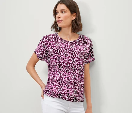 Frau posiert in einem bedruckten Blusenshirt, berry, und weißen Hosen.
