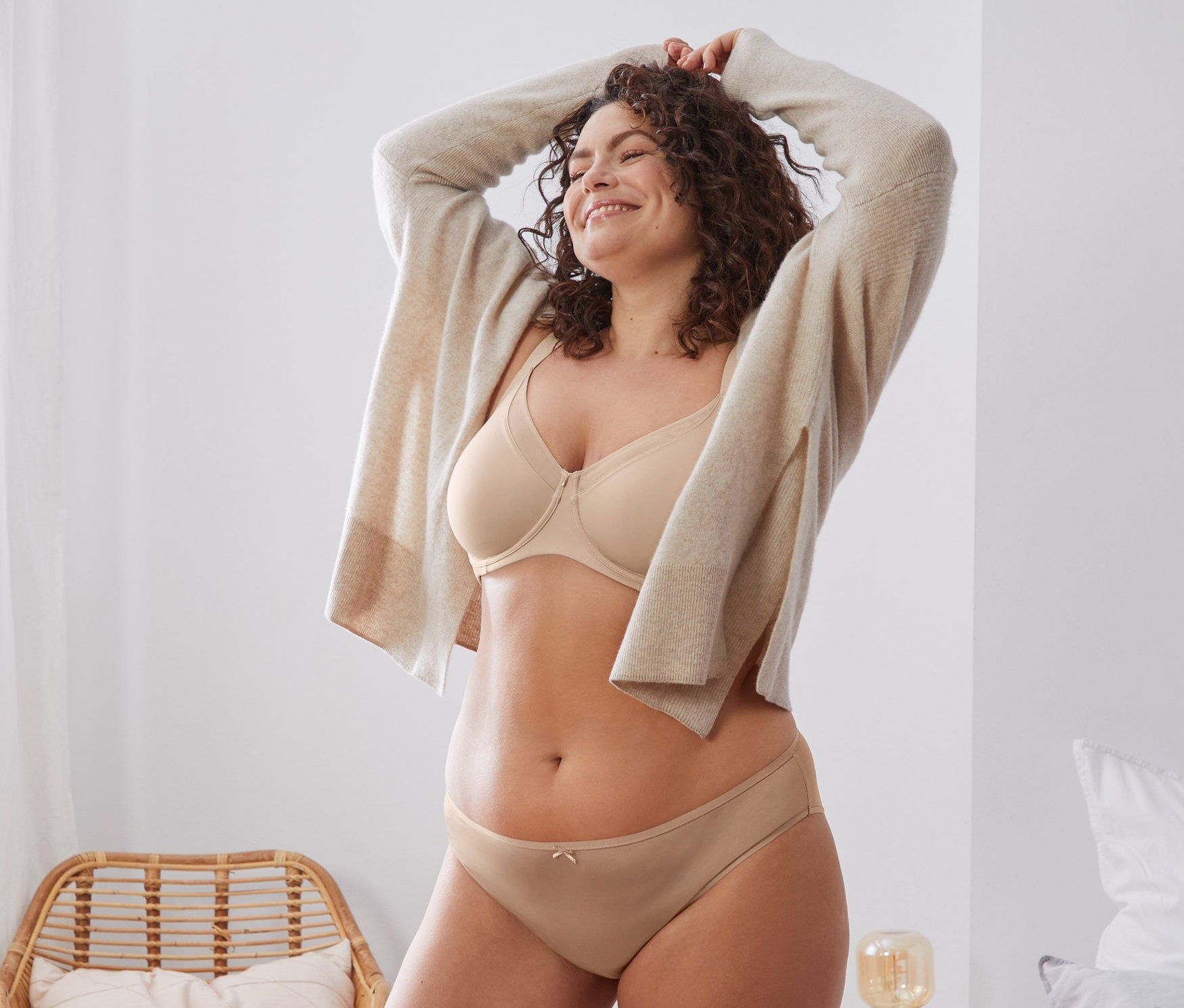 Frau lächelt und streckt die Hände über den Kopf. Sie trägt einen beigen Minimizer-BH und eine Strickjacke.