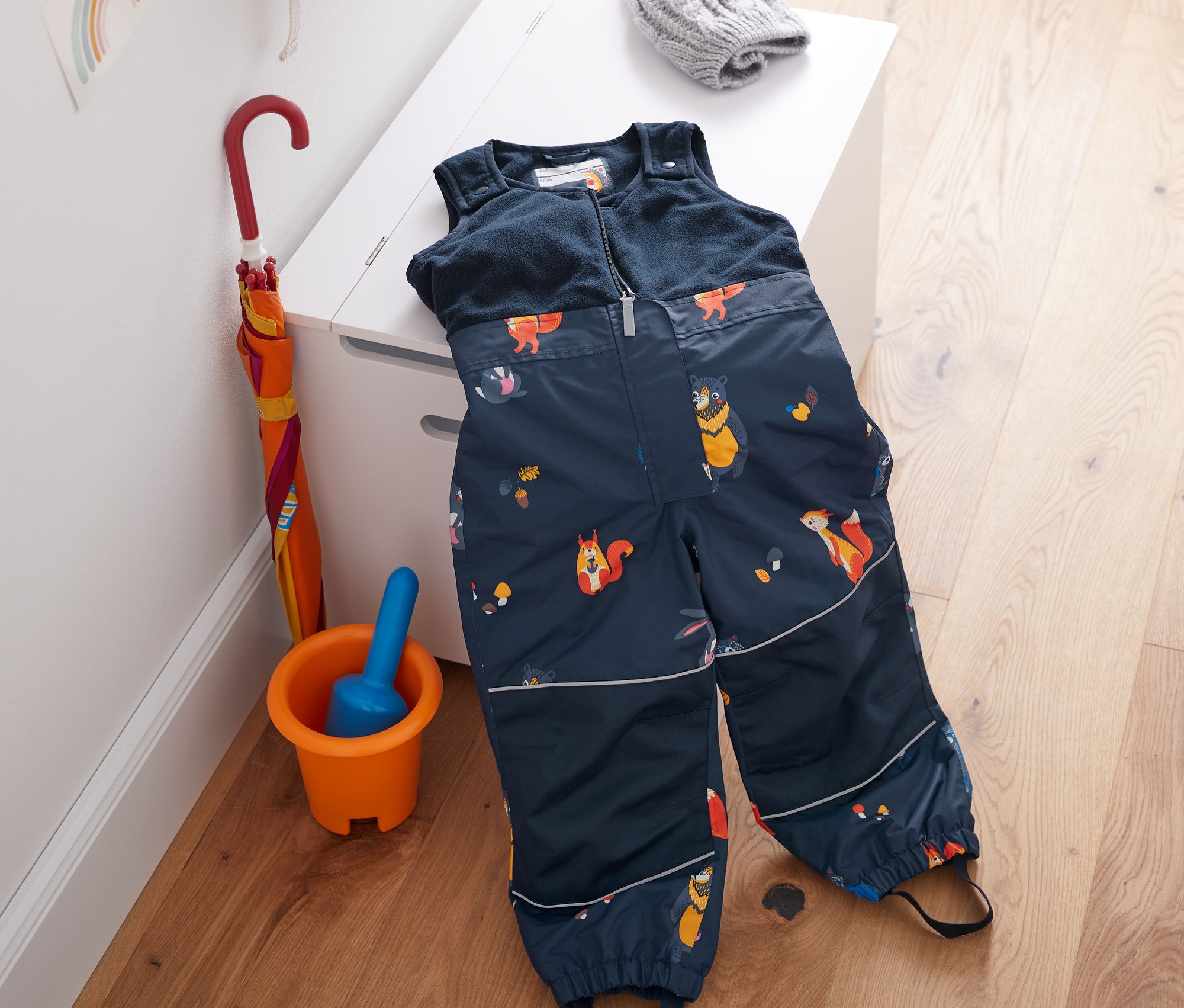 Thermo-Regenhose mit Fleecelatz aus recyceltem Material liegt auf einem weißen Schrank. Daneben Regenschirm, Eimer mit Schaufel und Rucksack.