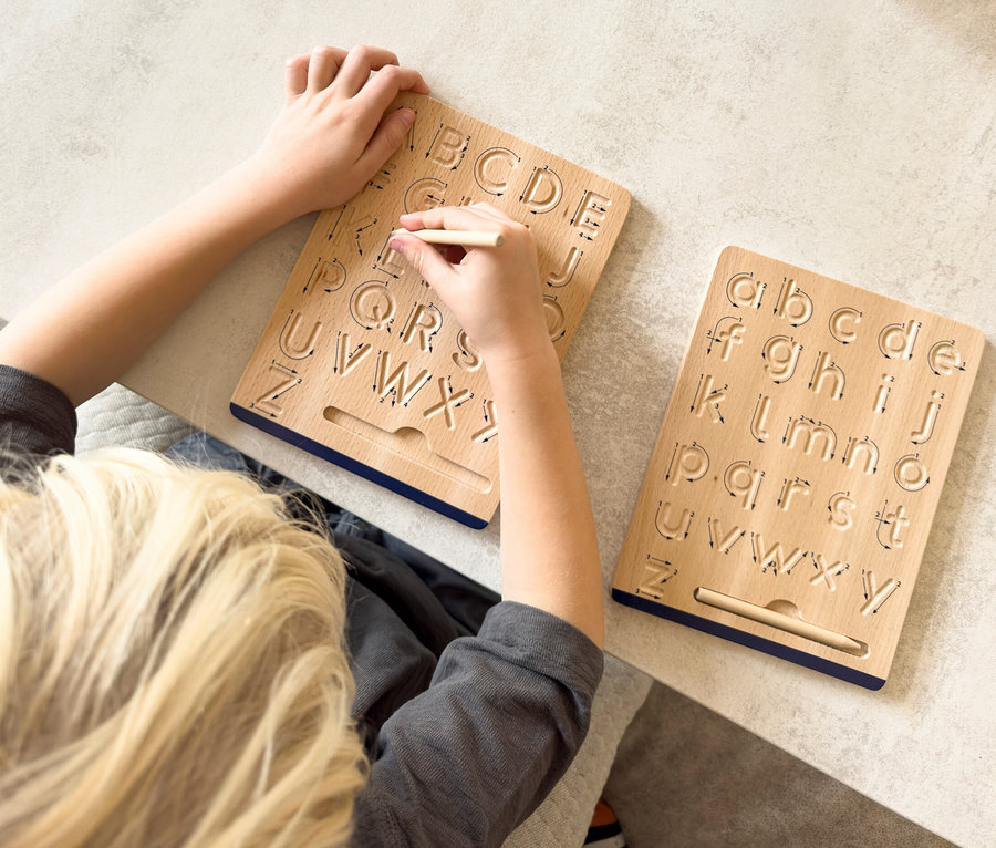 Kind schreibt mit Stift auf small foot Schreibtafel »Educate« mit gravierten Buchstaben. Auf dem Tisch liegen Papier und Stifte.