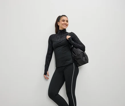 Lächelnde Frau in schwarzer Sportbekleidung mit einer Tasche über der Schulter.