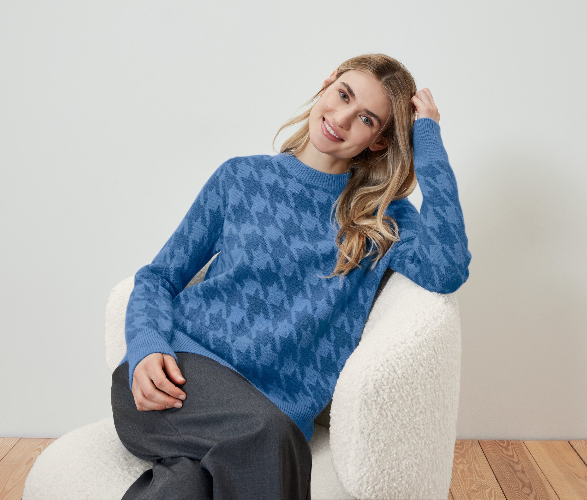 Blonde Frau sitzt in einem Sessel und trägt einen blauen Pullover mit Muster.
