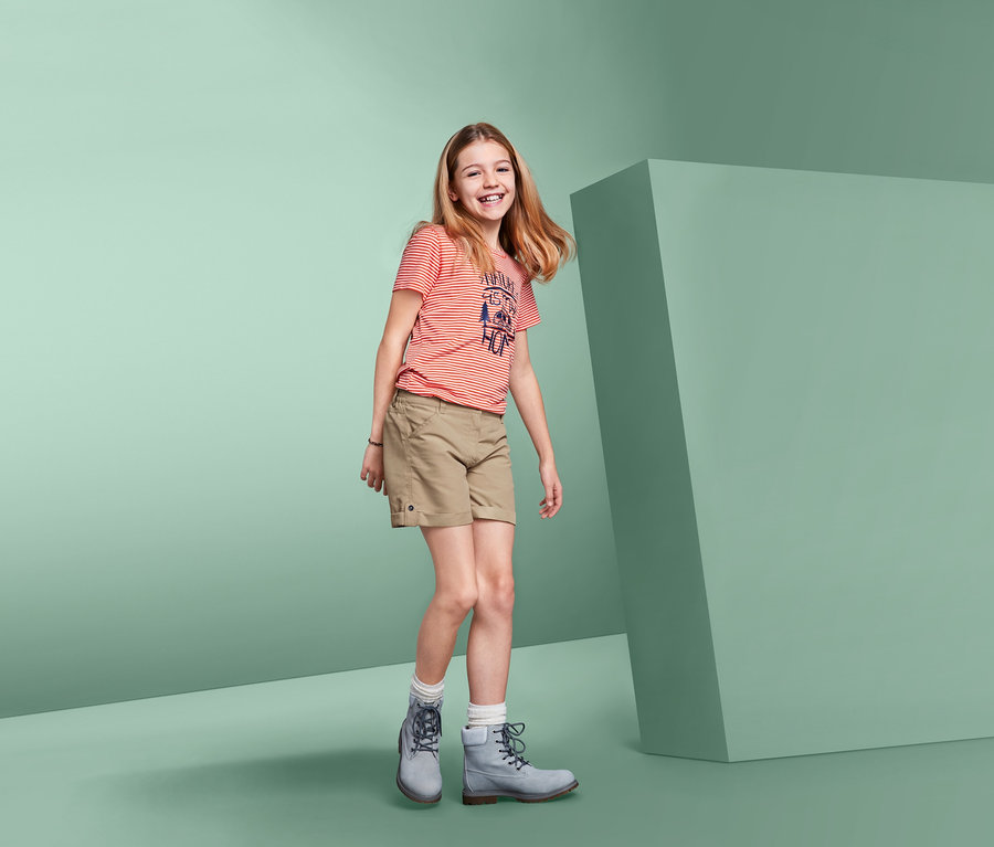 Lächelndes Mädchen posiert in beigen 2-in-1-Funktionsshorts, einem gestreiften T-Shirt und grauen Stiefeln.