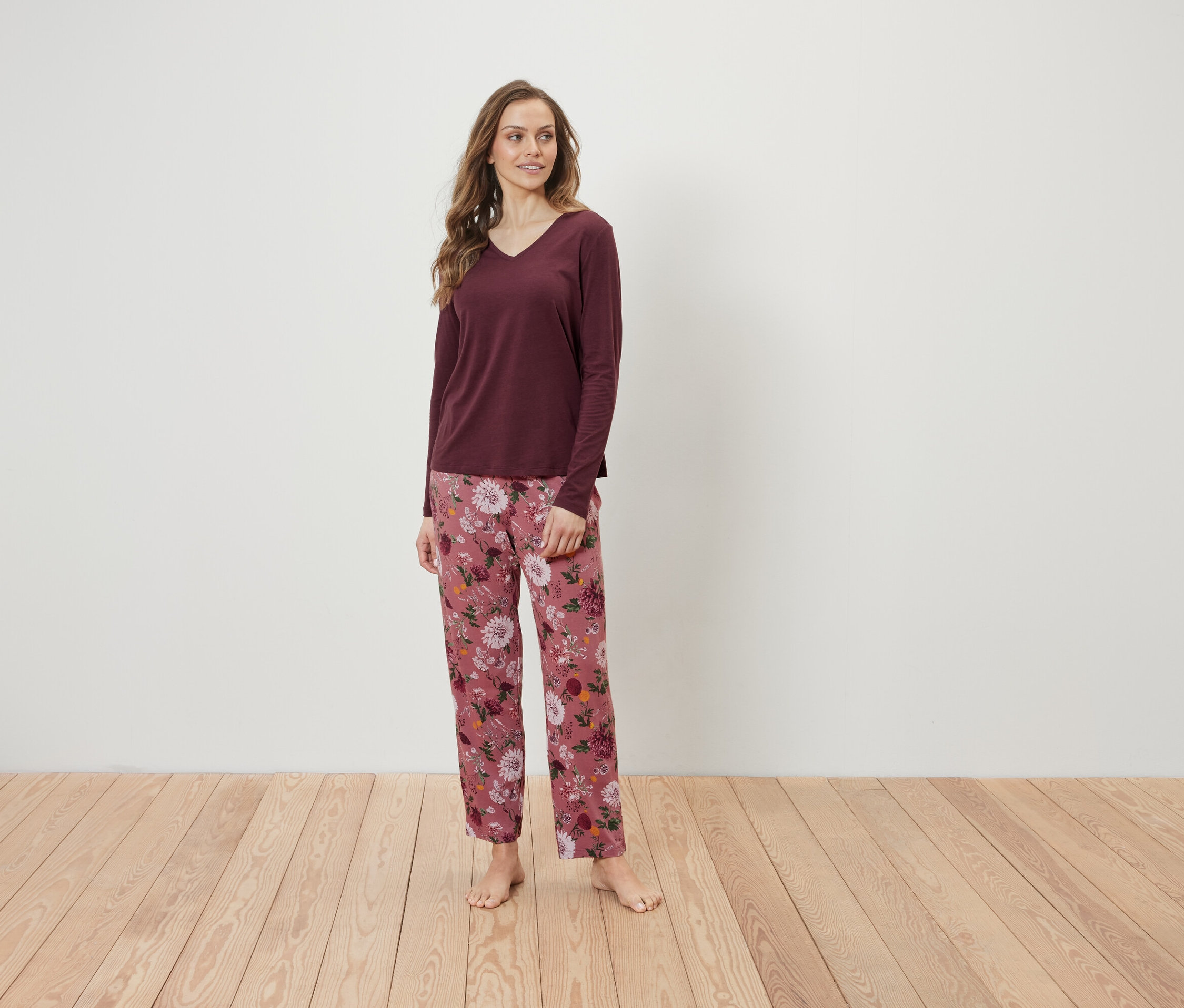 Eine Frau steht auf einem Holzboden und trägt ein weinrotes Langarmshirt und eine rosafarbene Blumenhose.