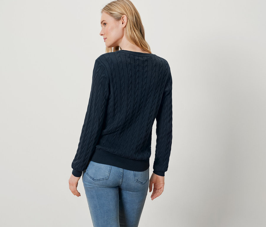 Frau mit blonden Haaren, bekleidet mit einem blauen Pullover und Jeans, steht mit dem Rücken zur Kamera.