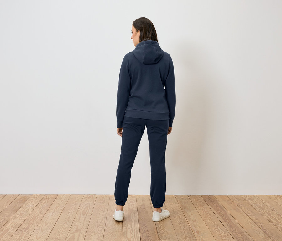 Frau von hinten in blauem Hoodie und Hose steht auf Holzboden vor einer weißen Wand.